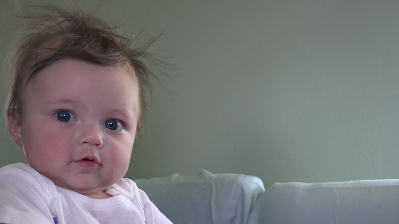 Baby sits near the center of a Westminster, Colorado indoor frame, looking toward camera with tousled hair, a white shirt, couch cushions, and a plain green wall visible.