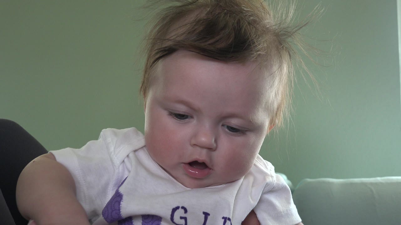 A baby in a white shirt looks forward near the camera in Westminster, Colorado, with a pale green wall and couch behind them.