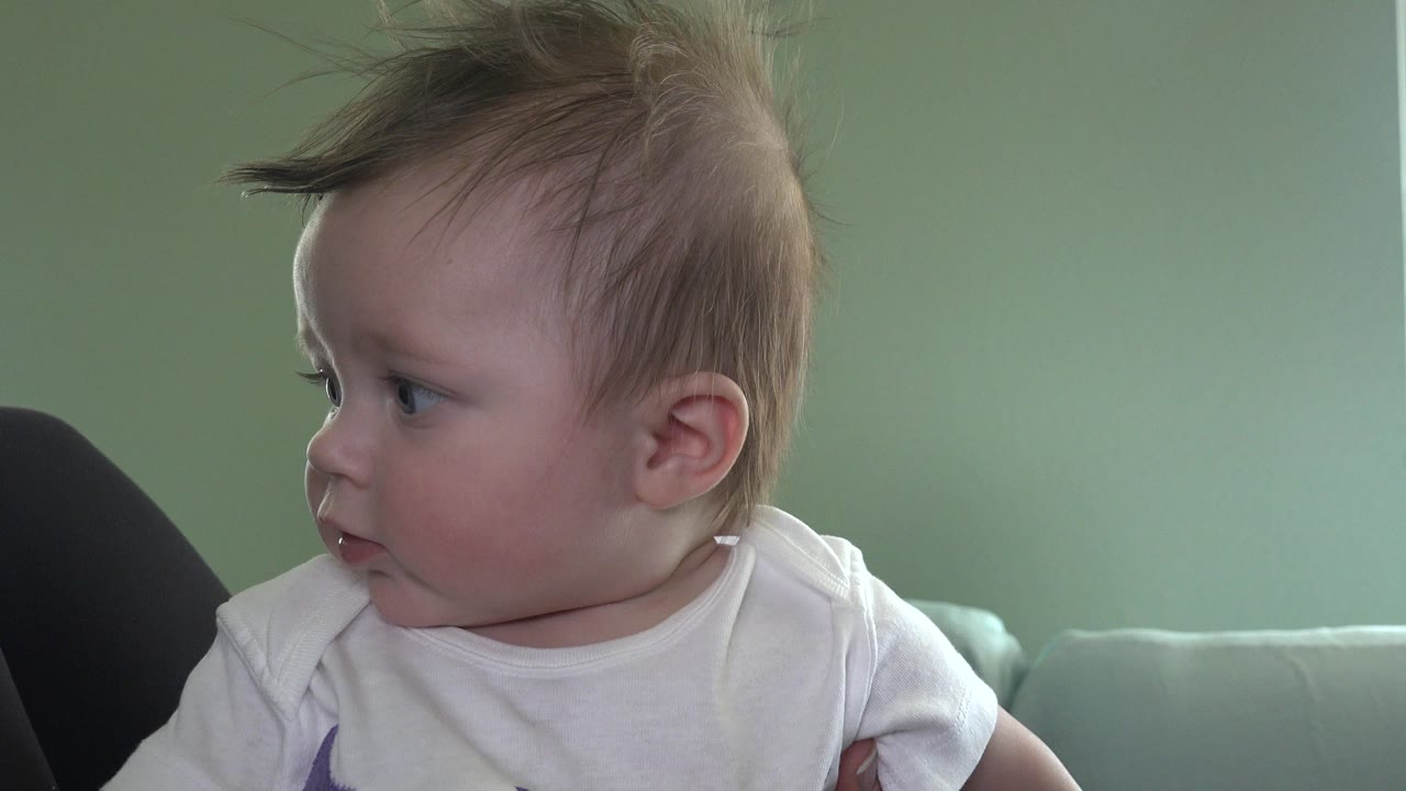 Baby in a white shirt looking left while being held indoors.