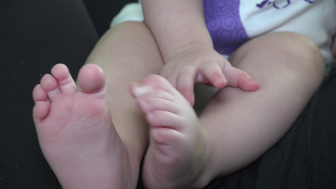 At 0:00, a baby in Westminster, Colorado sits on a dark couch with both bare feet close to the camera and hands resting near the legs.