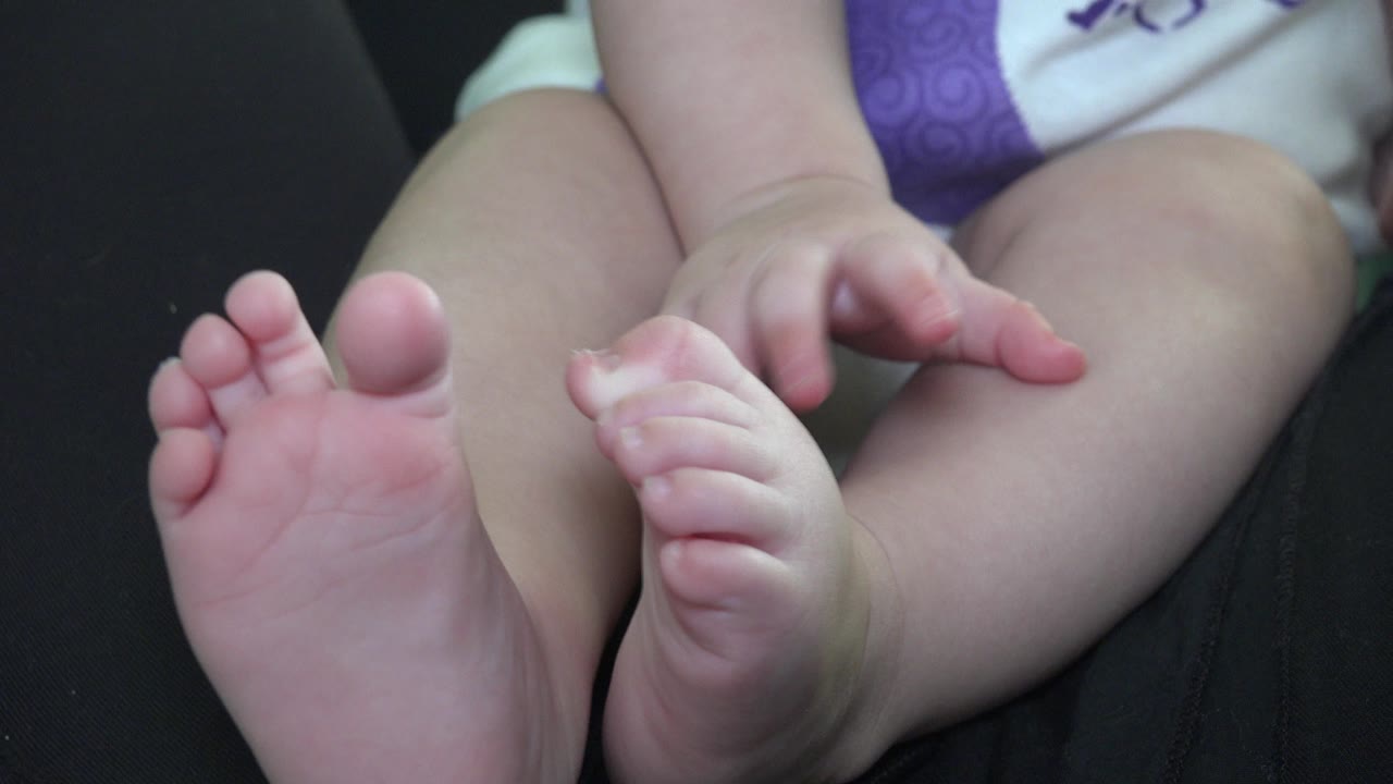 At 0:01, the baby's bare feet remain in the foreground on the dark couch, with one foot angled forward and small hands near the thighs.