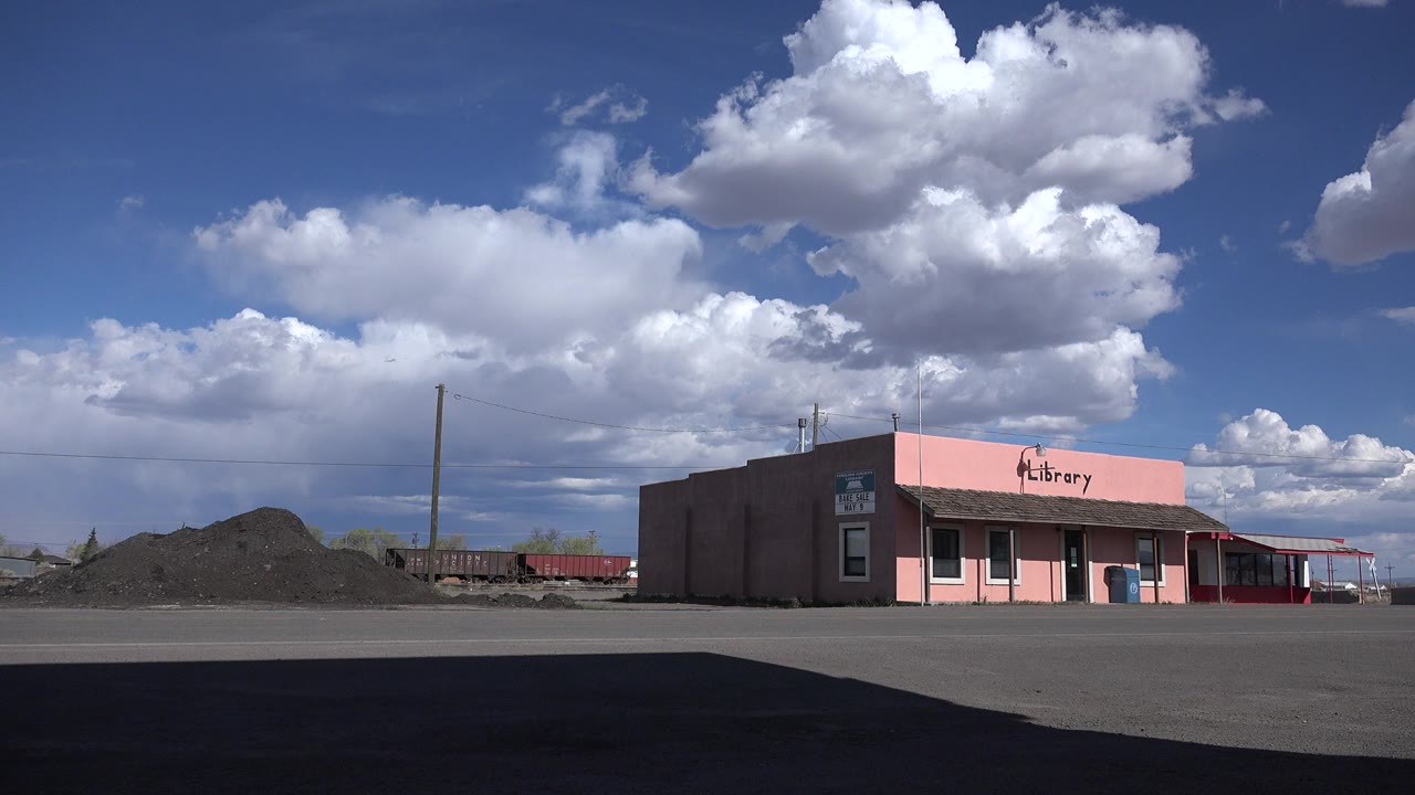 Wide Sterling, Colorado roadside view of a pink library building, gravel piles, utility poles, and a blue sky with large clouds, with no visible people.