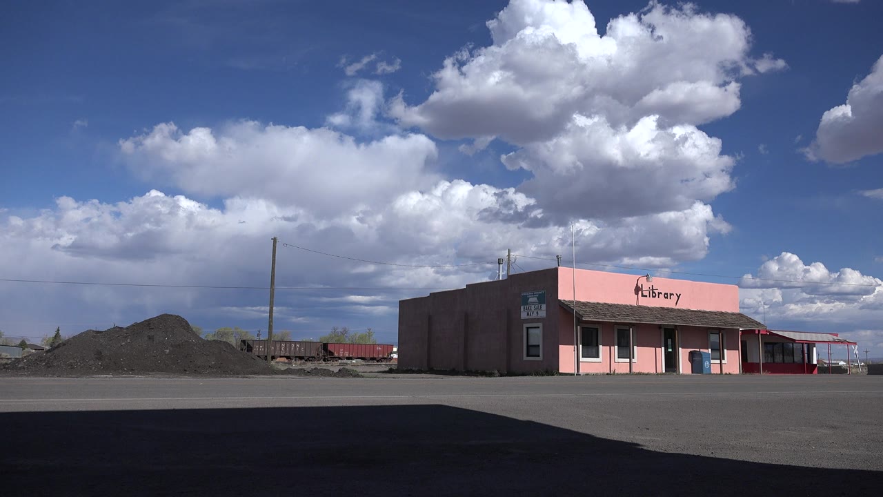 Wide view of a pink library building in Sterling, Colorado, with dark pavement in the foreground, material piles to the left, large white clouds overhead, and no visible people.