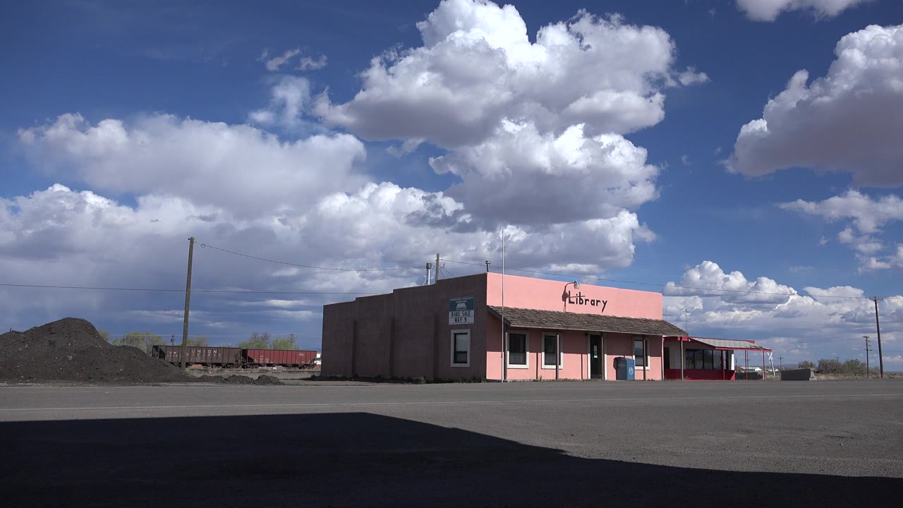 Sterling, Colorado frame showing the pink low-rise library slightly right of center, a dark foreground shadow on the asphalt, a gravel mound to the left, and no visible people.