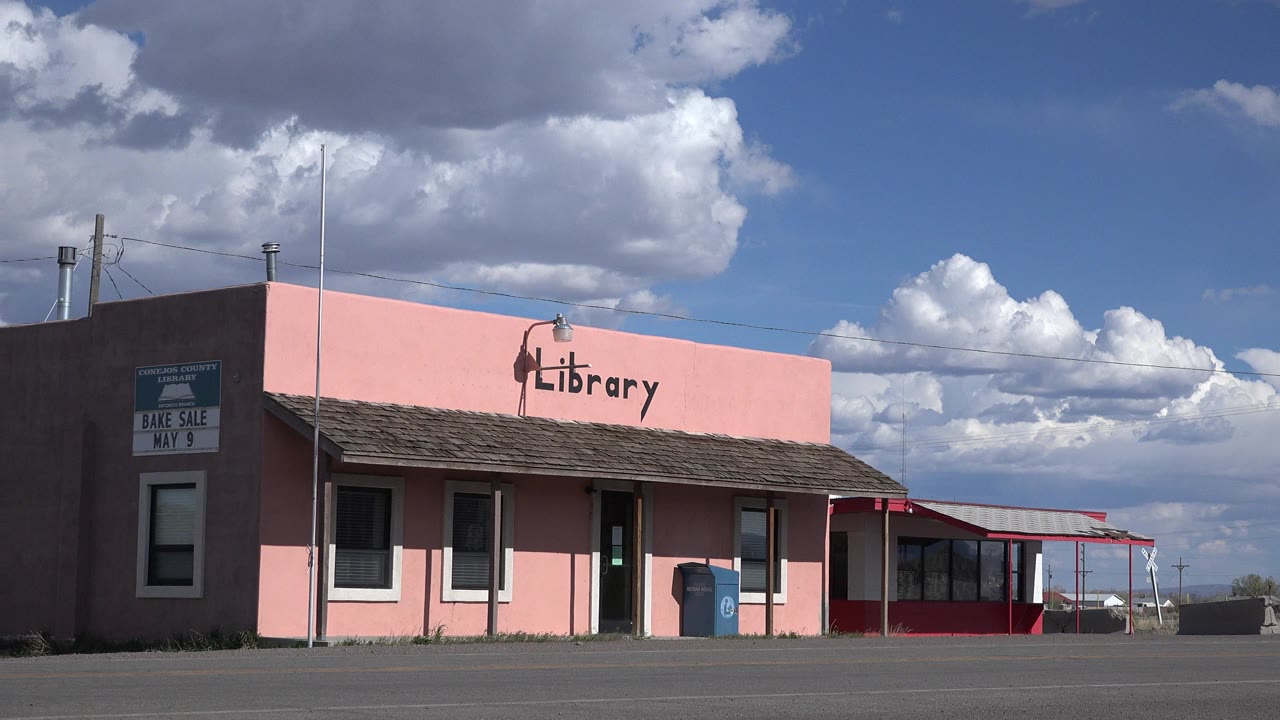 Wide view of a pink library building in Sterling, Colorado, seen across pavement under large white clouds with no visible people.