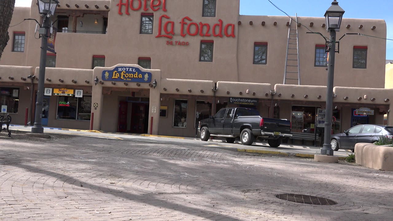 Opening frame of the Hotel La Fonda facade in Taos, New Mexico, with red wall signage, a blue entrance sign, parked vehicles, street lamps, and no visible people.