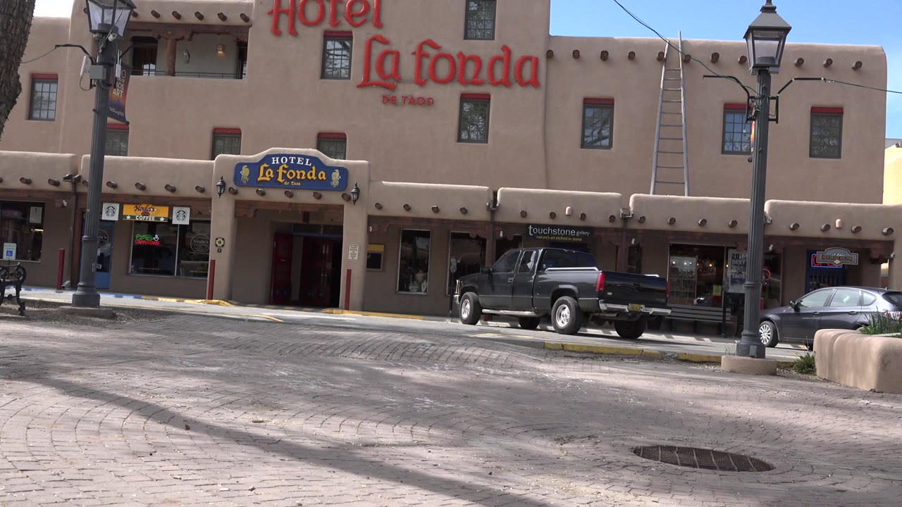 Second frame holds on the Taos hotel facade with the entrance centered lower in frame, a ladder on the upper wall, parked cars along the curb, and no visible people.