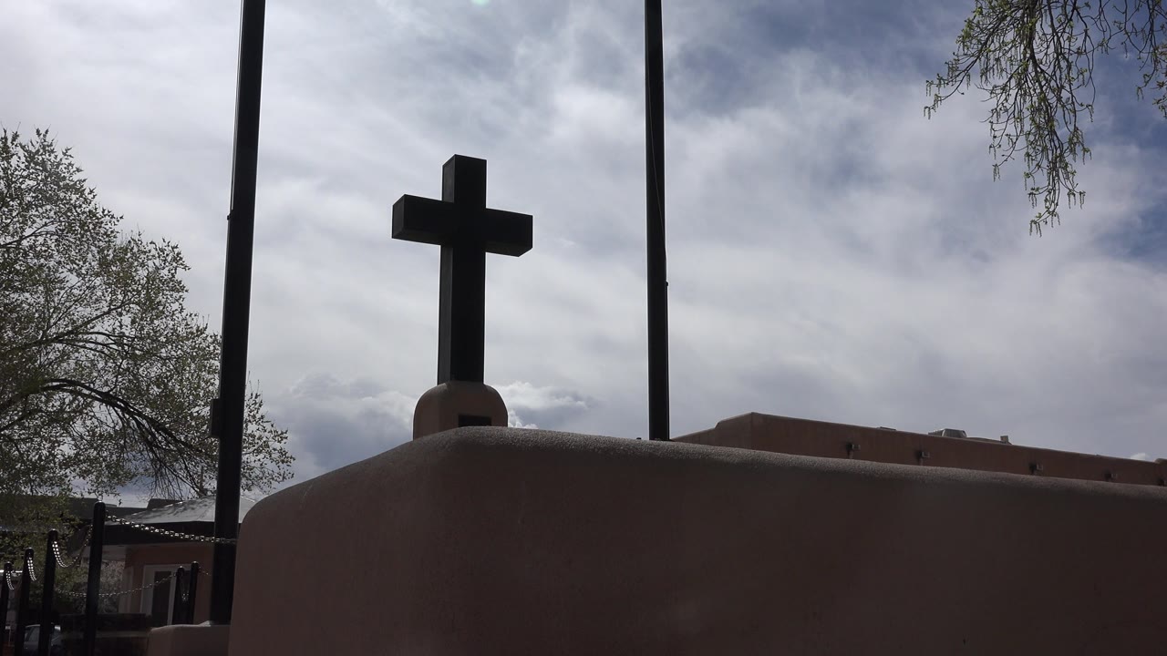 At 0:01, the cross remains centered above the rounded adobe roofline in Taos New Mexico, with tree branches at the edges, cloud cover overhead, and no visible people.