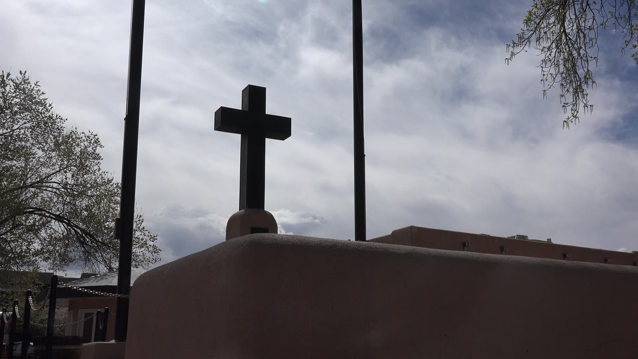 At the start of the Taos New Mexico clip, a dark cross stands above a rounded adobe-toned church parapet with cloudy sky, tree edges, and no visible people.