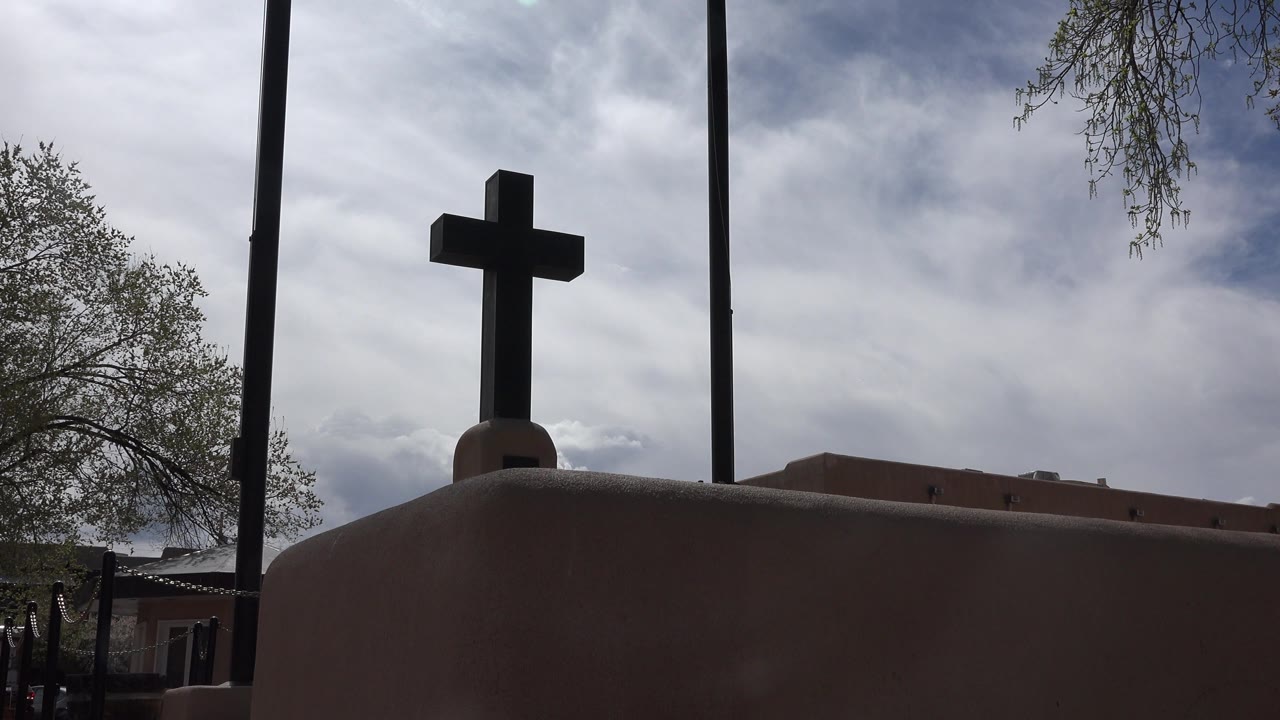 Opening frame of a dark cross above an adobe-toned facade in Taos New Mexico, with tree branches at the sides, tall poles behind it, cloudy sky, and no visible people.