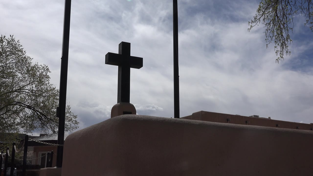 Dark church cross in Taos New Mexico above an adobe-colored foreground wall, with trees at the edges, vertical poles, cloudy sky, and no visible people.