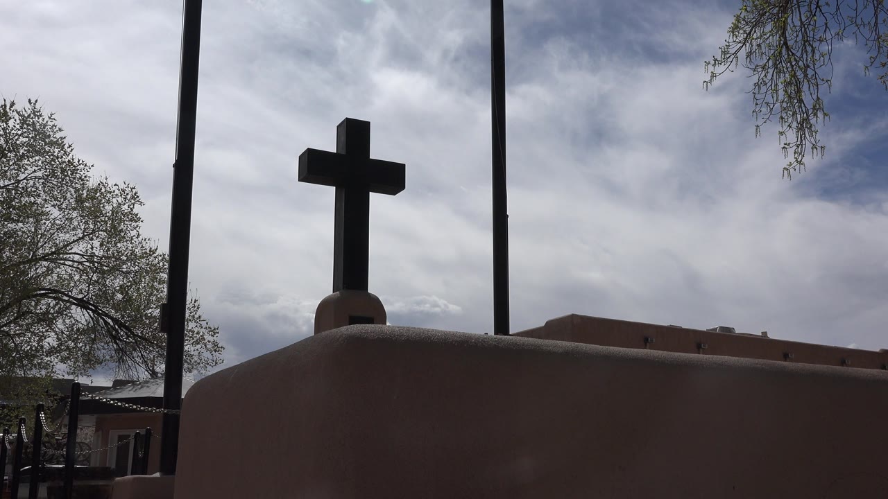 Dark cross above an adobe-colored wall under a cloudy sky.