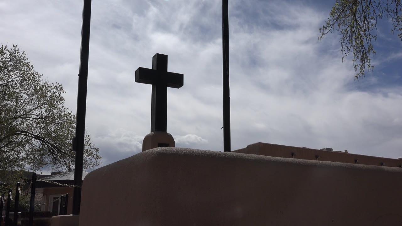 Opening frame of a dark church cross in Taos, New Mexico, seen above a rounded adobe-colored wall with two vertical poles and no visible people.