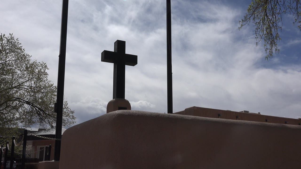 Dark cross above a tan exterior wall under cloudy sky.