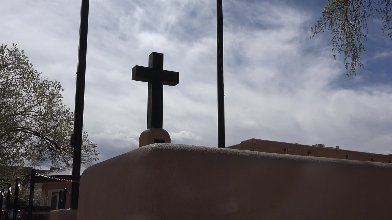 At 0:00, a dark cross stands above an adobe-colored church roof in Taos New Mexico, with tall poles, tree branches, clouded blue sky, and no visible people.