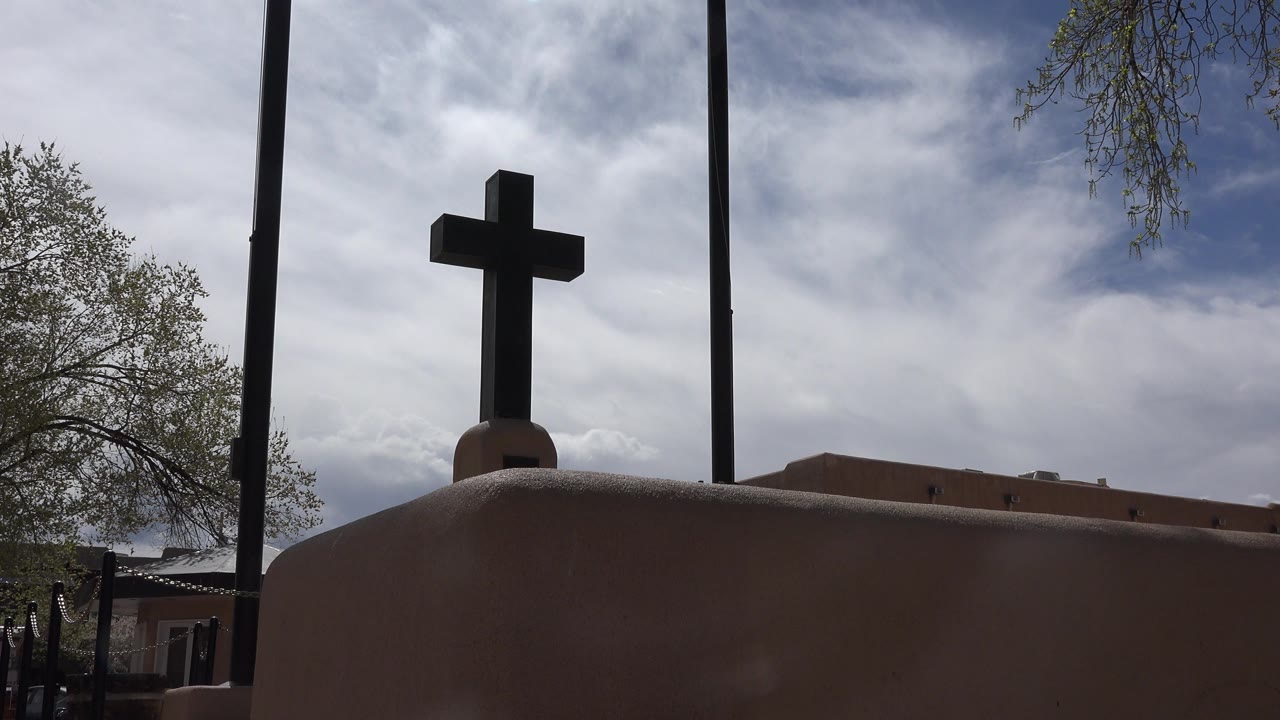 Dark cross above an adobe-colored wall under a cloudy sky.