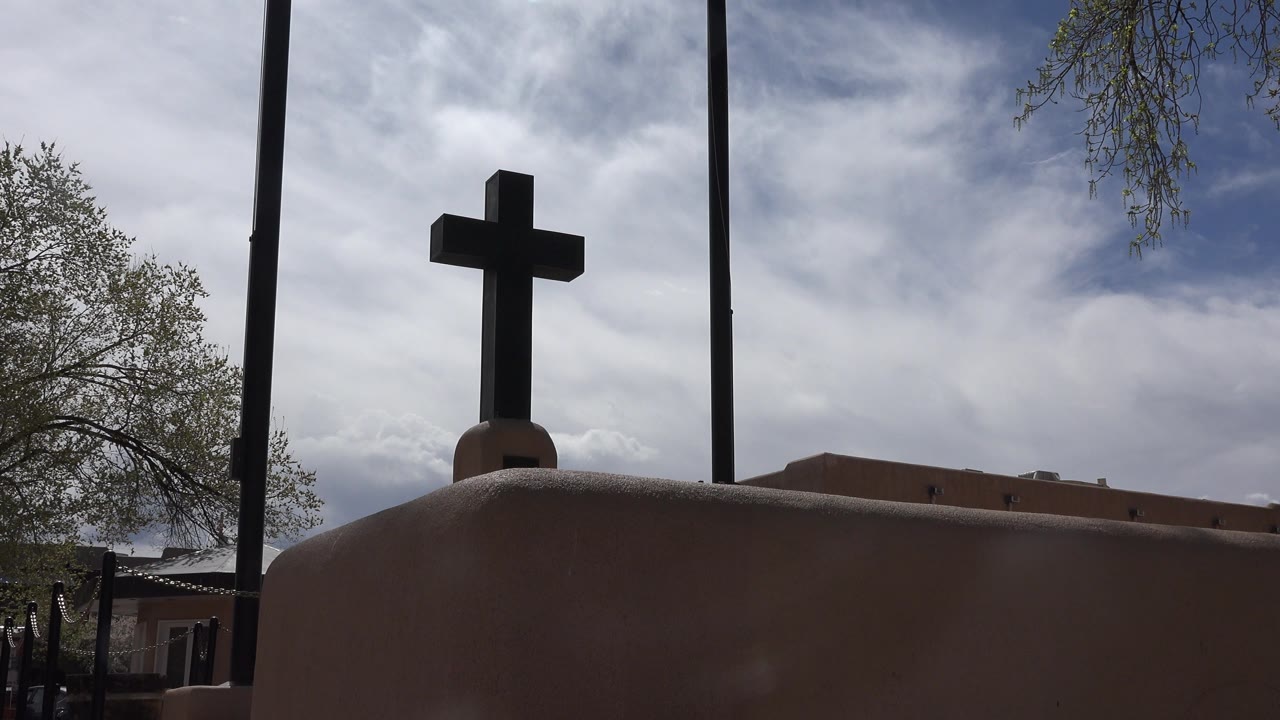 Opening frame in Taos New Mexico showing a dark cross on a rounded adobe-colored tomb or wall, with cloudy blue sky, trees, poles, low buildings, and no visible people.