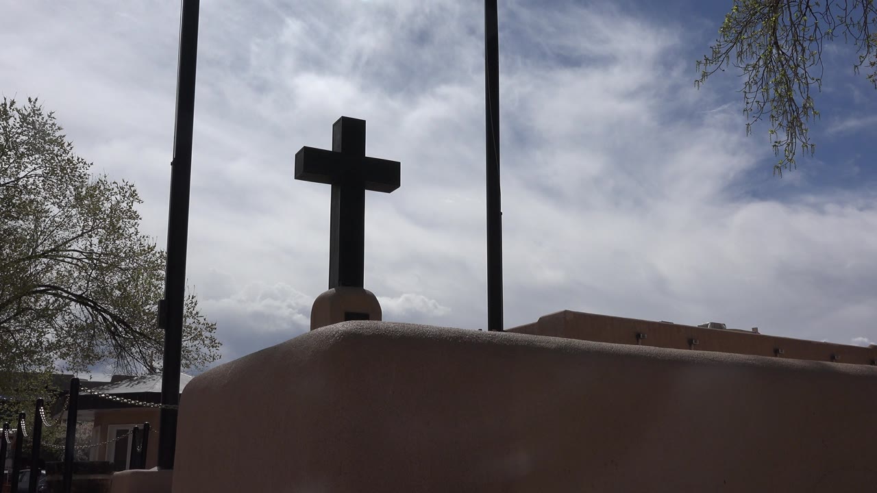 Dark cross above an adobe-colored wall under a cloudy blue sky.