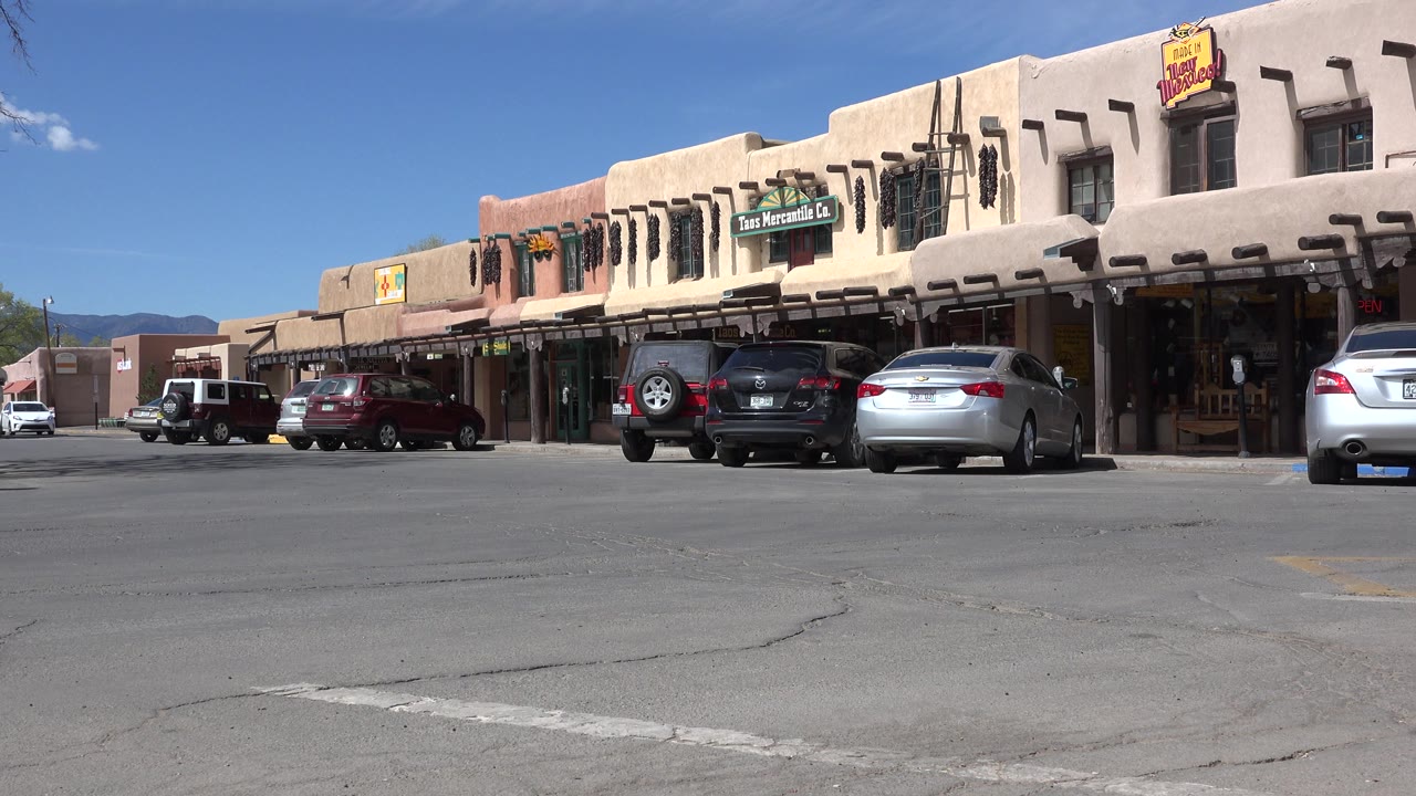 Second frame showing the same Taos New Mexico storefront row, with a white vehicle at left, several parked cars near the shops, and no visible people.