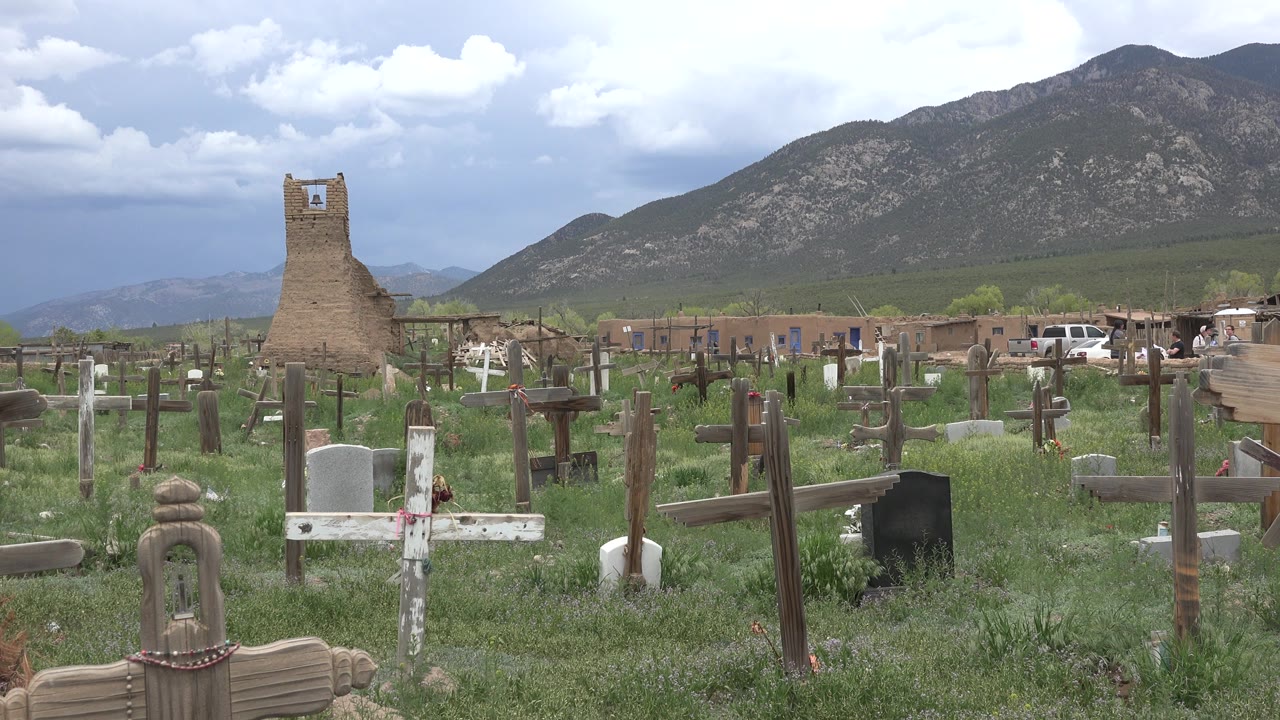 Wooden crosses and grave markers fill a grassy cemetery in Taos New Mexico, with an adobe-like tower, cloudy sky, and no visible people.