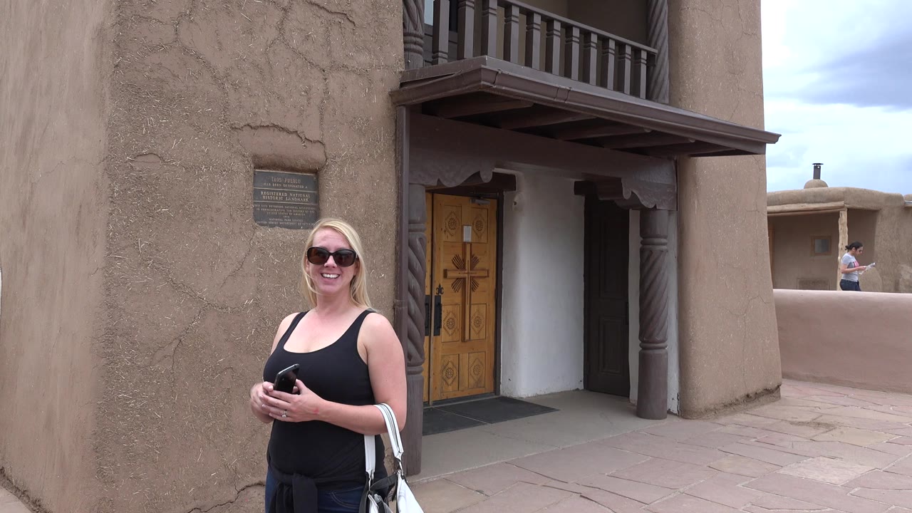 Blonde woman standing beside an adobe religious building entrance.