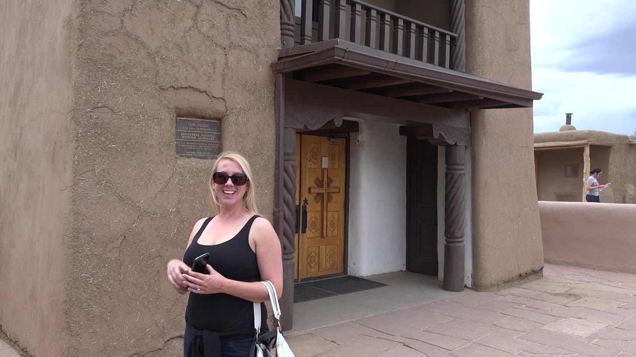 Smiling blonde tourist in sunglasses faces the camera near carved wooden doors and tan adobe walls at the Taos New Mexico religious site.