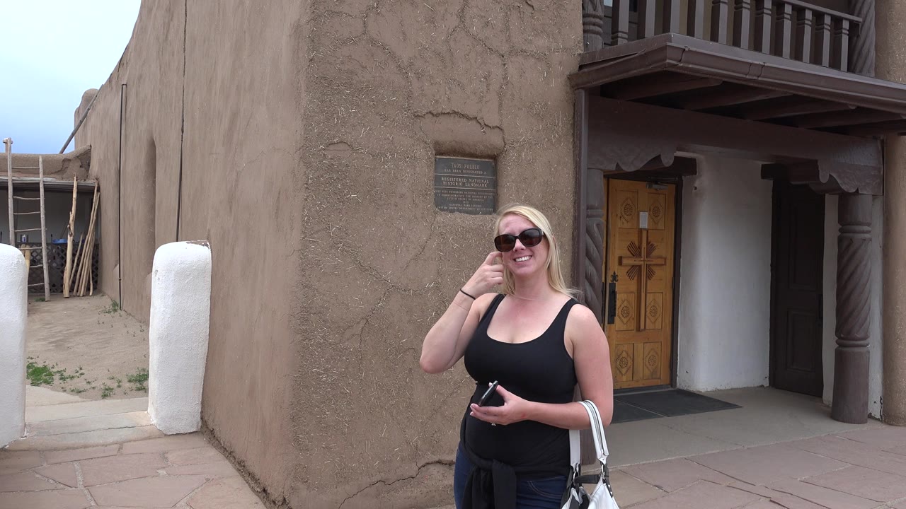 Blonde woman in sunglasses standing by a tan adobe-style building.