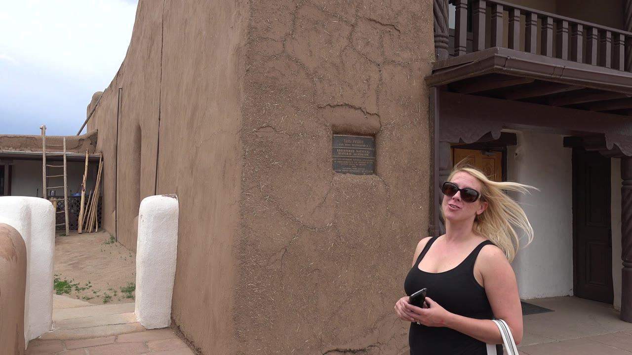 Smiling woman in sunglasses standing by an adobe-style building entrance.