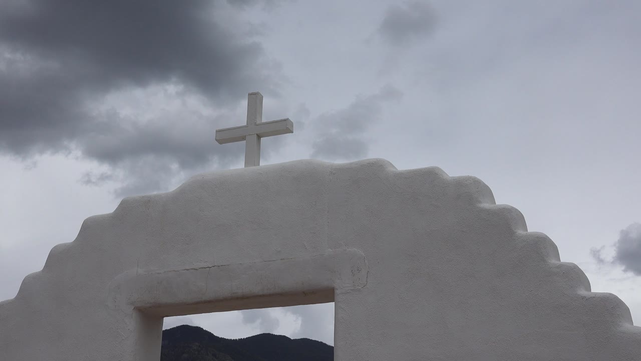 Second frame holds on the cross above the white stepped archway in Taos New Mexico, with a darker cloud mass moving across the upper sky and no visible people.