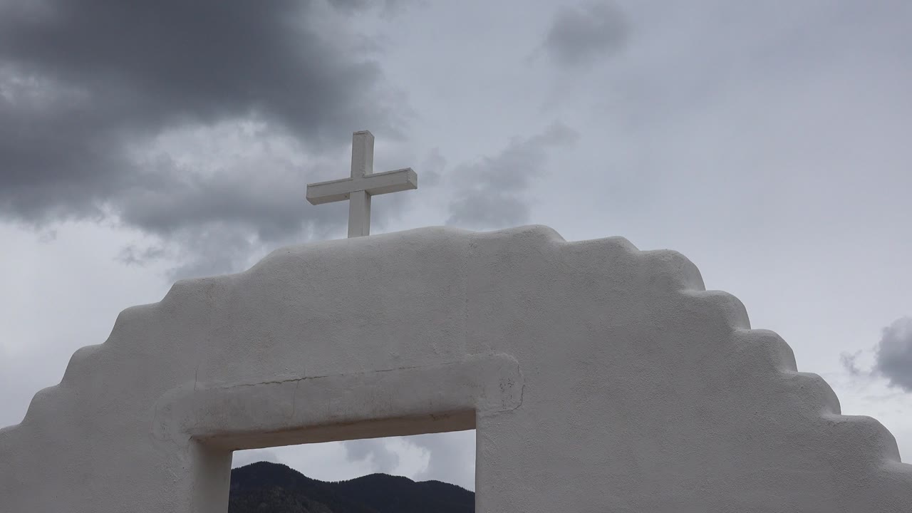 Opening frame of a white stepped archway in Taos, New Mexico, with a cross above the roofline, gray clouds overhead, dark hills through the opening, and no visible people.