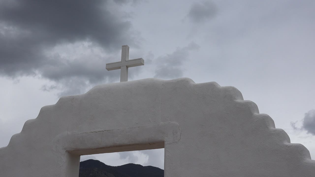 At one second, the cross remains near the top of the white Taos archway while a darker cloud mass sits above it and no visible people appear in frame.