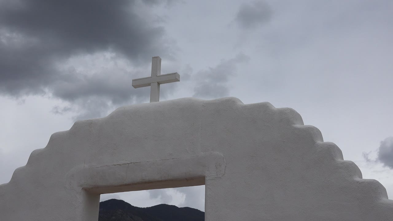 Close upward view of a white archway and cross below clouds.