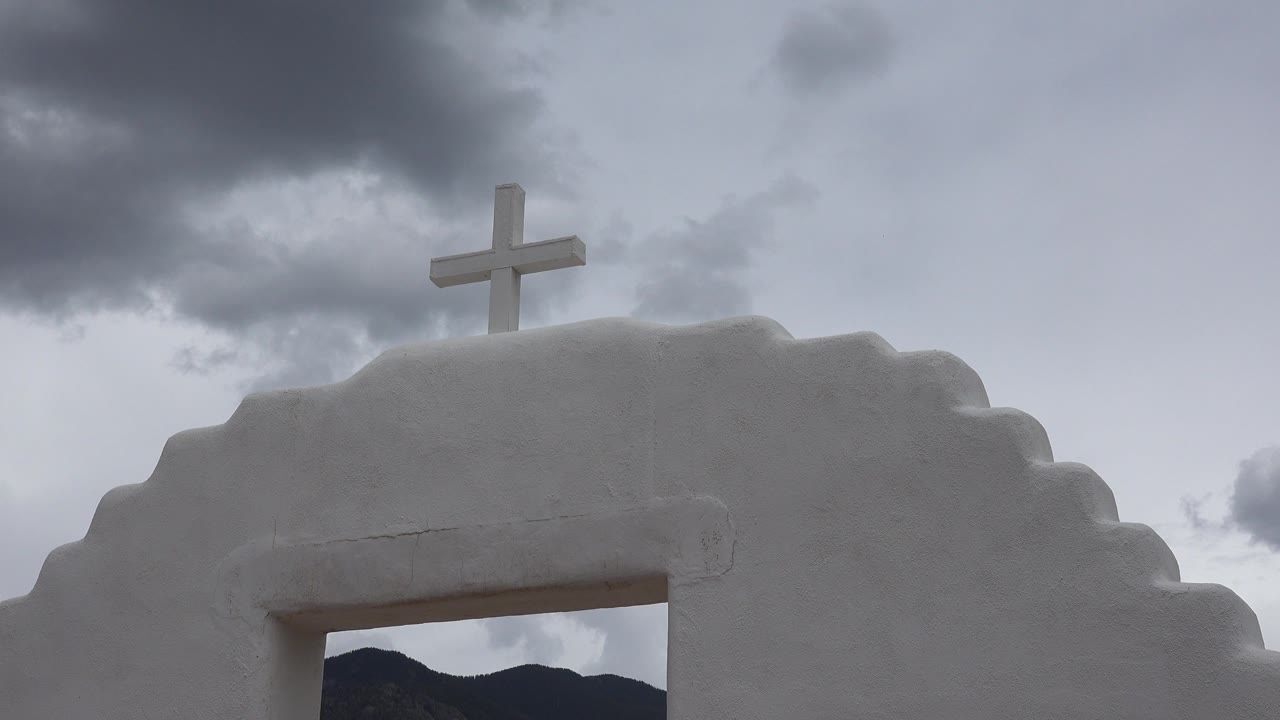 At 0:00, a white stepped arch or roofline in Taos New Mexico sits below a pale cross, with gray clouds above and no visible people.