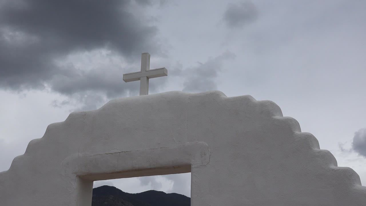 White cross rises above a stepped white building arch in Taos New Mexico, with gray clouds overhead and no visible people.