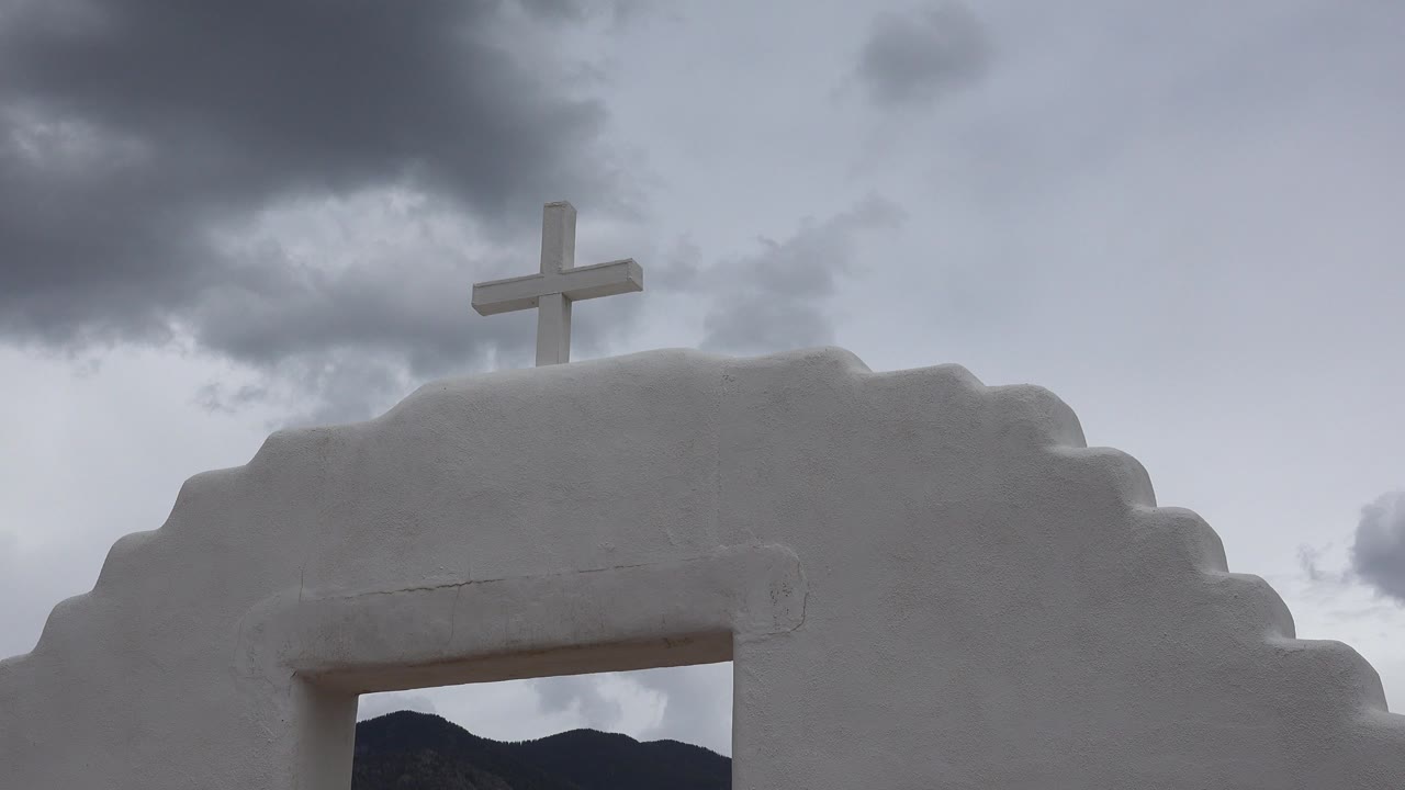 The cross remains near the upper center above the white arch, with dark cloud cover filling the Taos New Mexico sky and no visible people.