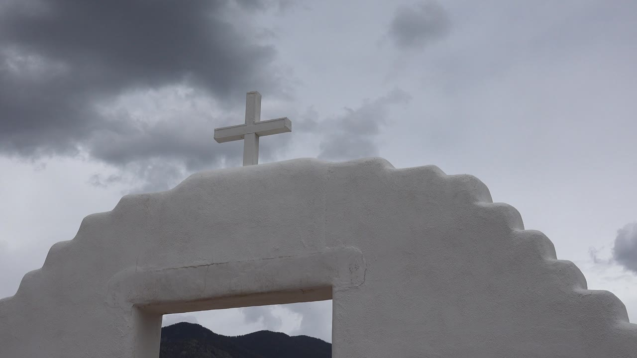 A white cross stands on the top edge of the white arch in Taos New Mexico, with a darker cloud mass at upper left and no visible people.
