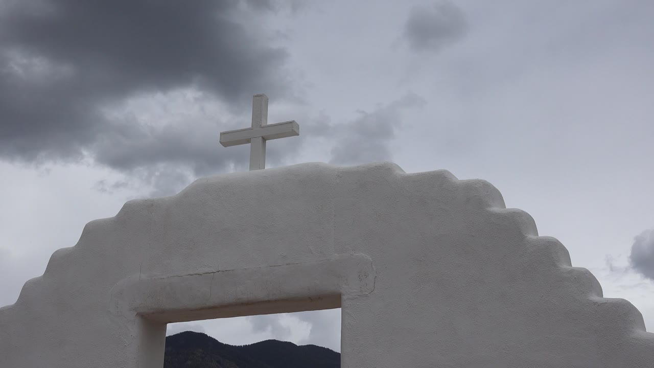 The white chapel cross in Taos New Mexico rises into a pale gray sky while a darker cloud mass hangs above the roofline, with no visible people.