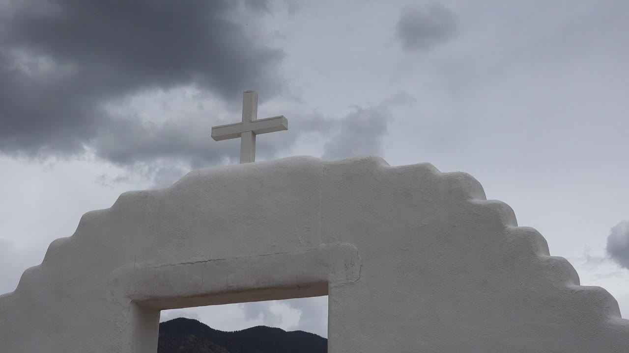 Opening frame of a white stepped church facade in Taos New Mexico with a cross near the roofline, gray clouds overhead, dark hills through the lower opening, and no visible people.