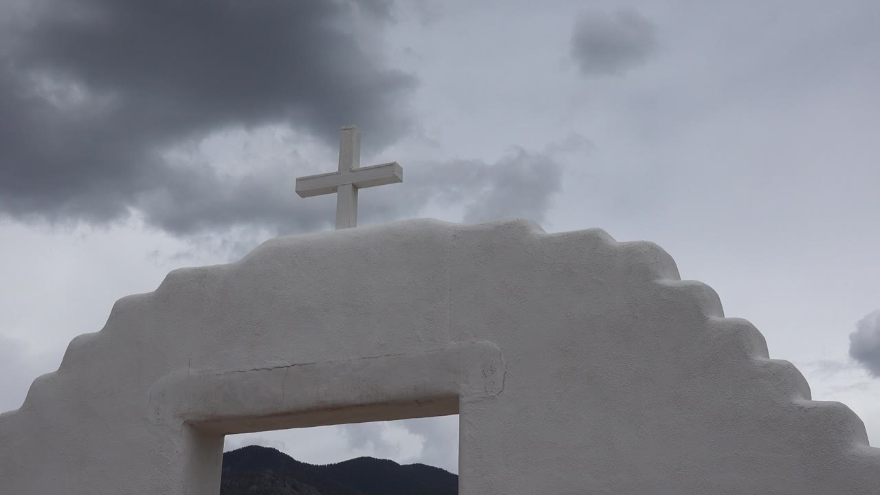 Opening frame of a white adobe-style facade in Taos New Mexico with a small cross near the top, gray clouds above, and no visible people.