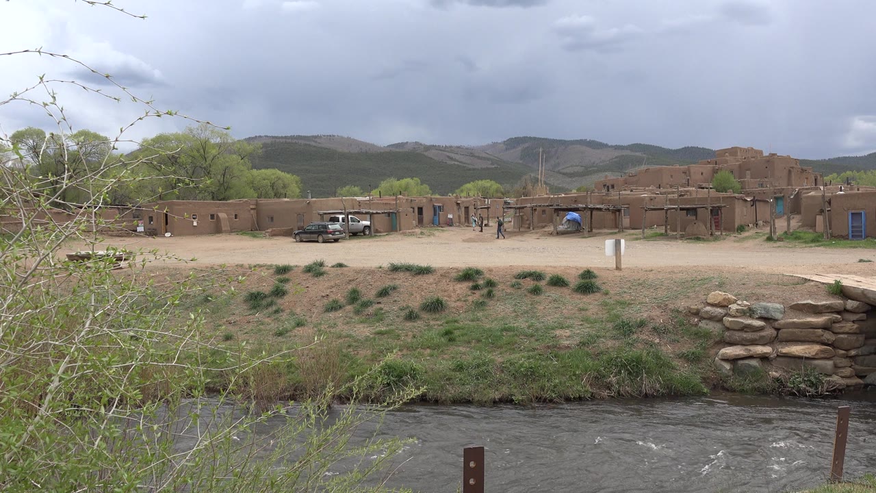 Wide Taos, New Mexico exterior with a river at the bottom of frame, sparse shrubs, tan adobe-style buildings, parked vehicles, distant people, and mountains beneath gray clouds.