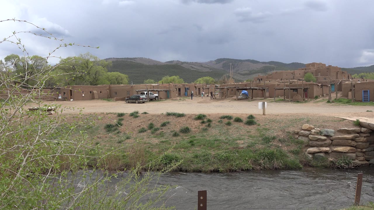 River in front of roadside adobe-style homes under cloudy sky.