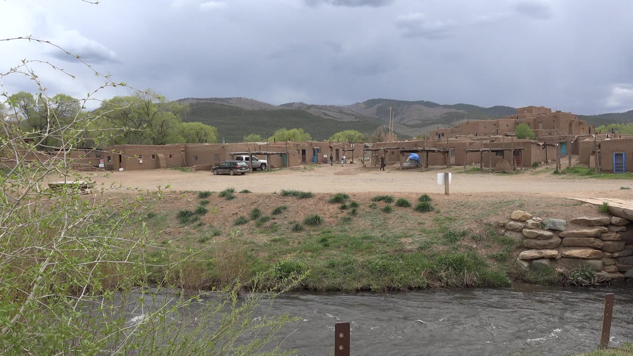 Stream in foreground with sandy bank, shrubs, adobe-style buildings, parked vehicles, hills, and cloudy sky.