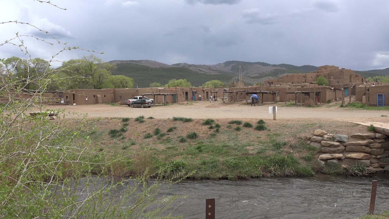 At 0:00 in Taos, New Mexico, dark river water runs below brush, with adobe-style buildings, a parked pickup, low hills, and heavy gray clouds beyond, with no visible people.