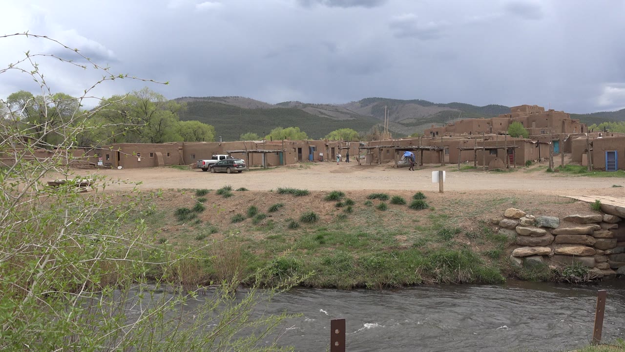 At 0:02 in Taos, New Mexico, rippled river water crosses the lower frame, with scrub, a stone edge, earthen buildings, a pickup truck, and dark cloud cover in the distance, with no visible people.
