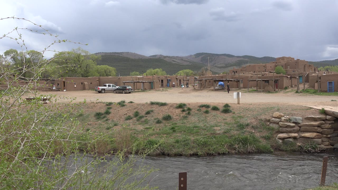 Opening frame of the Taos, New Mexico clip shows a fast river in the foreground, brush at left, adobe-style buildings, parked cars, distant people, and green hills beneath gray clouds.