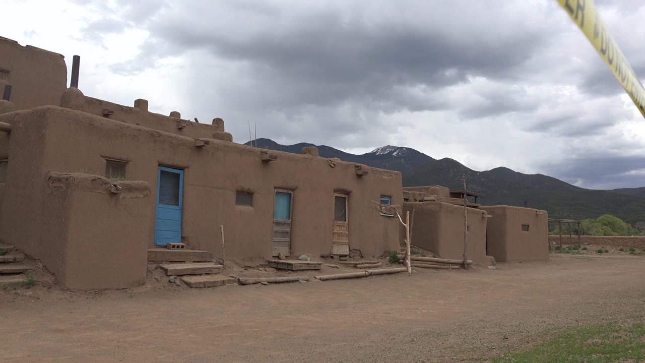 Adobe-style homestead buildings with blue doors under dark clouds.