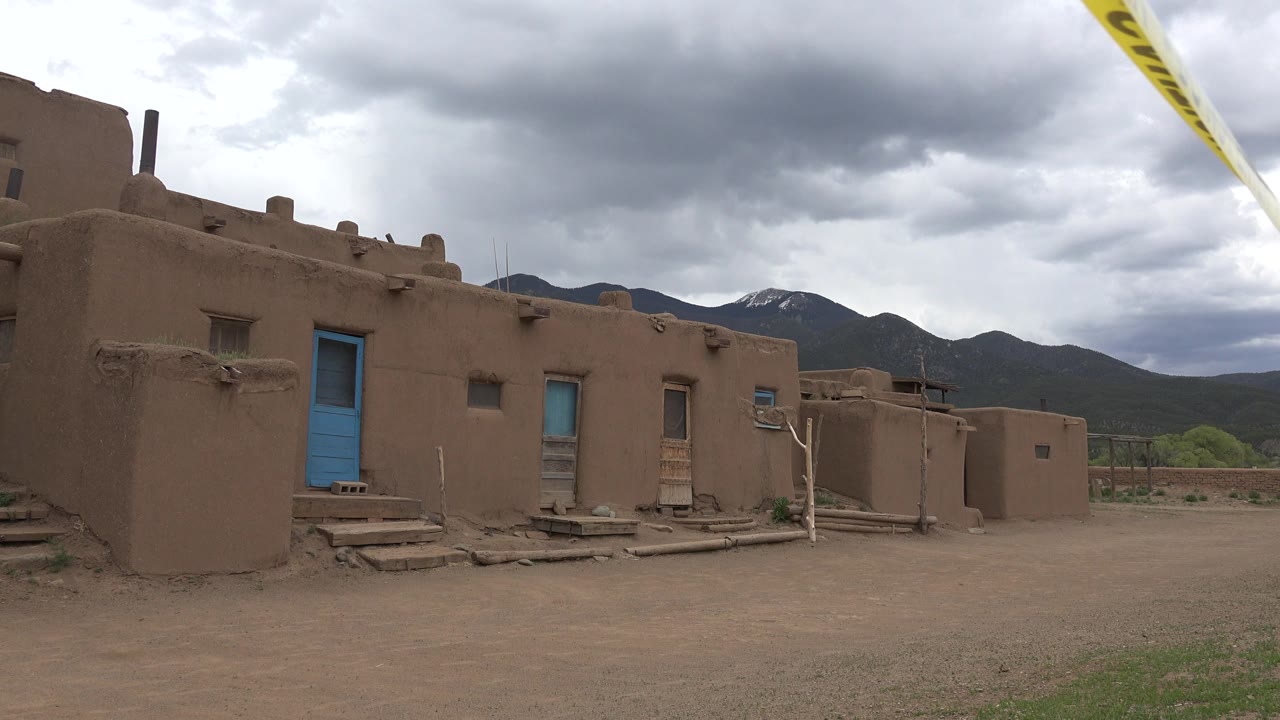 Taos, New Mexico homestead wall and blue doorway seen from the dirt yard, with mountains and heavy clouds in the background and no visible people.