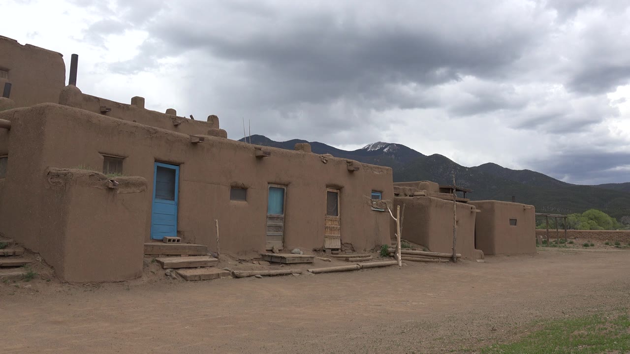 Adobe-style buildings with blue doors below cloudy mountains.