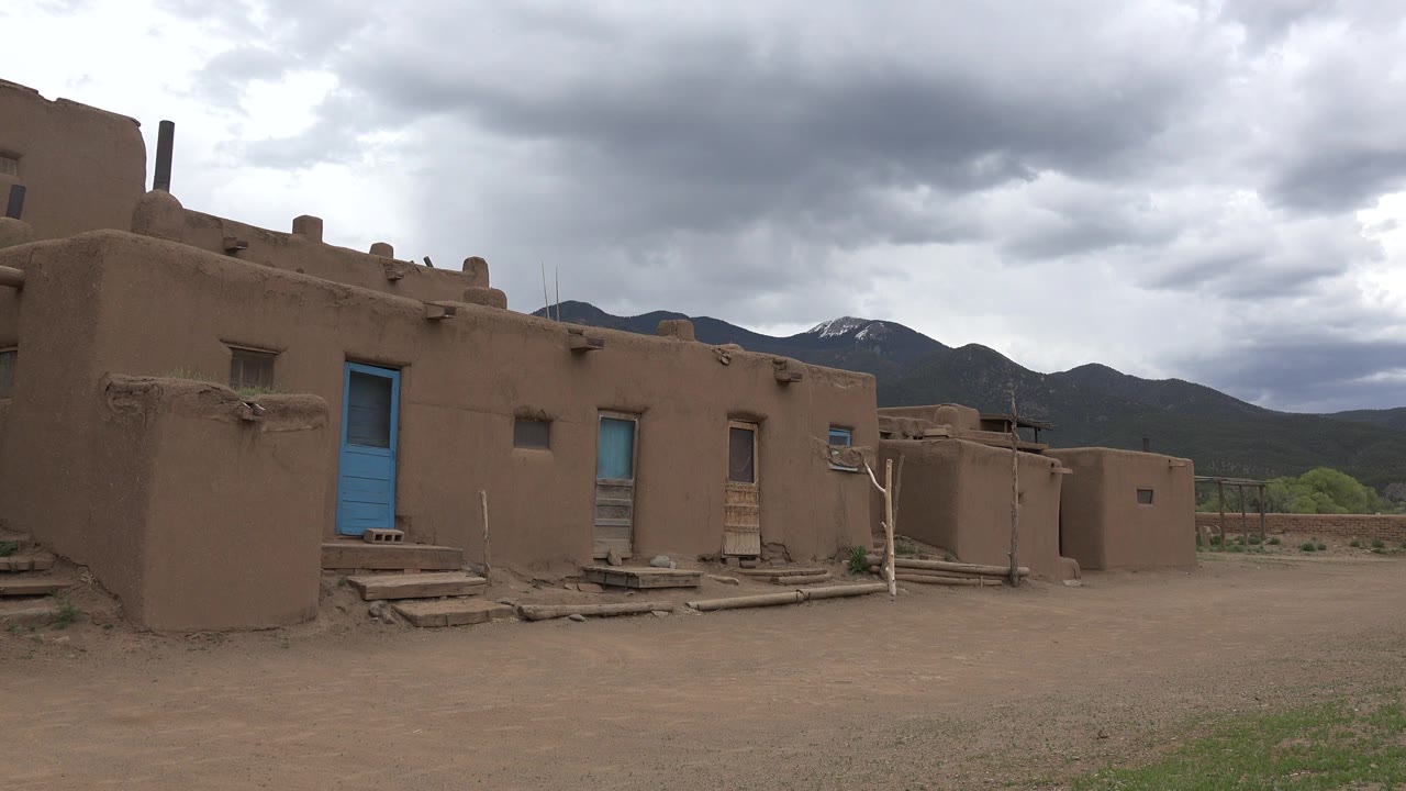 Taos New Mexico adobe building with blue doors at the left of frame, a dirt yard in front, mountains and gray clouds behind, and no visible people.