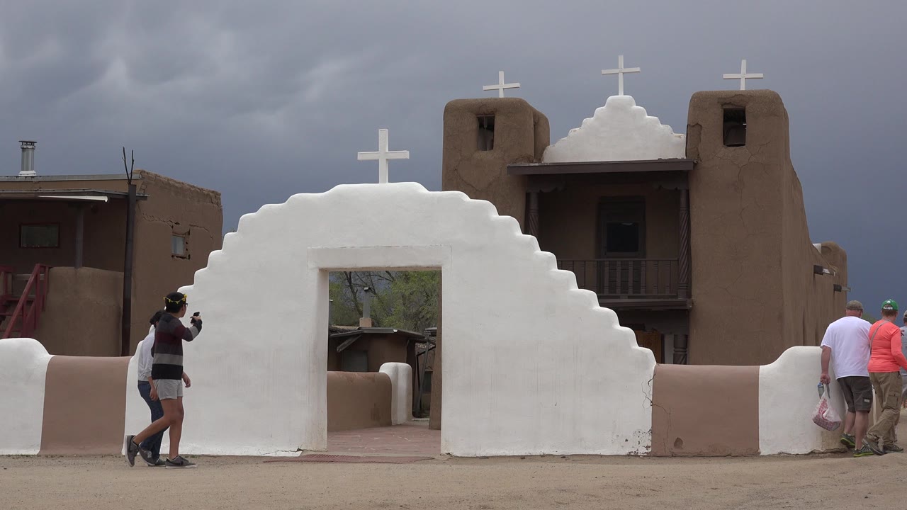 Two people walking beside a white church wall under dark clouds.